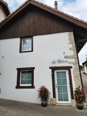 Un edificio con fachada blanca, detalles en marrón y un letrero que dice 'Au Chateau'. Tiene dos ventanas con contraventanas y dos macetas con flores rosadas.