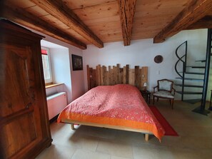 Un dormitorio con techo de madera, una cama con colcha rojo anaranjado, un armario de madera, una silla, una mesita, un cuadro en la pared y una escalera de caracol.