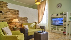 A living room with a green armchair, a sofa, a coffee table, a TV, a bookshelf, and a stone wall. - Anesis Studio Old Corfu Town (Corfu)