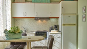 A compact kitchen with white cabinets, a built-in oven, a refrigerator, and a dining area with a table and two chairs. - Anesis Studio Old Corfu Town (Corfu)