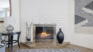 A room with a white brick fireplace, a modern table with a lamp, a patterned rug, and a large abstract painting on the wall.