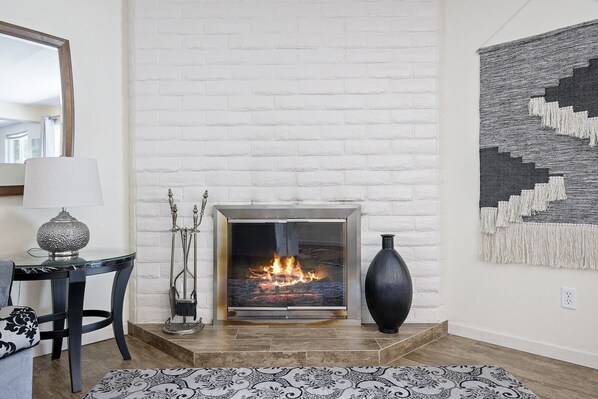Una habitación con chimenea de ladrillo blanco, una mesa moderna con lámpara, alfombra estampada y un cuadro abstracto grande en la pared.