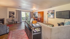 A spacious living room with a fireplace, a dining area, and a kitchen. There is a large window providing natural light and a flat-screen TV mounted on the wall.