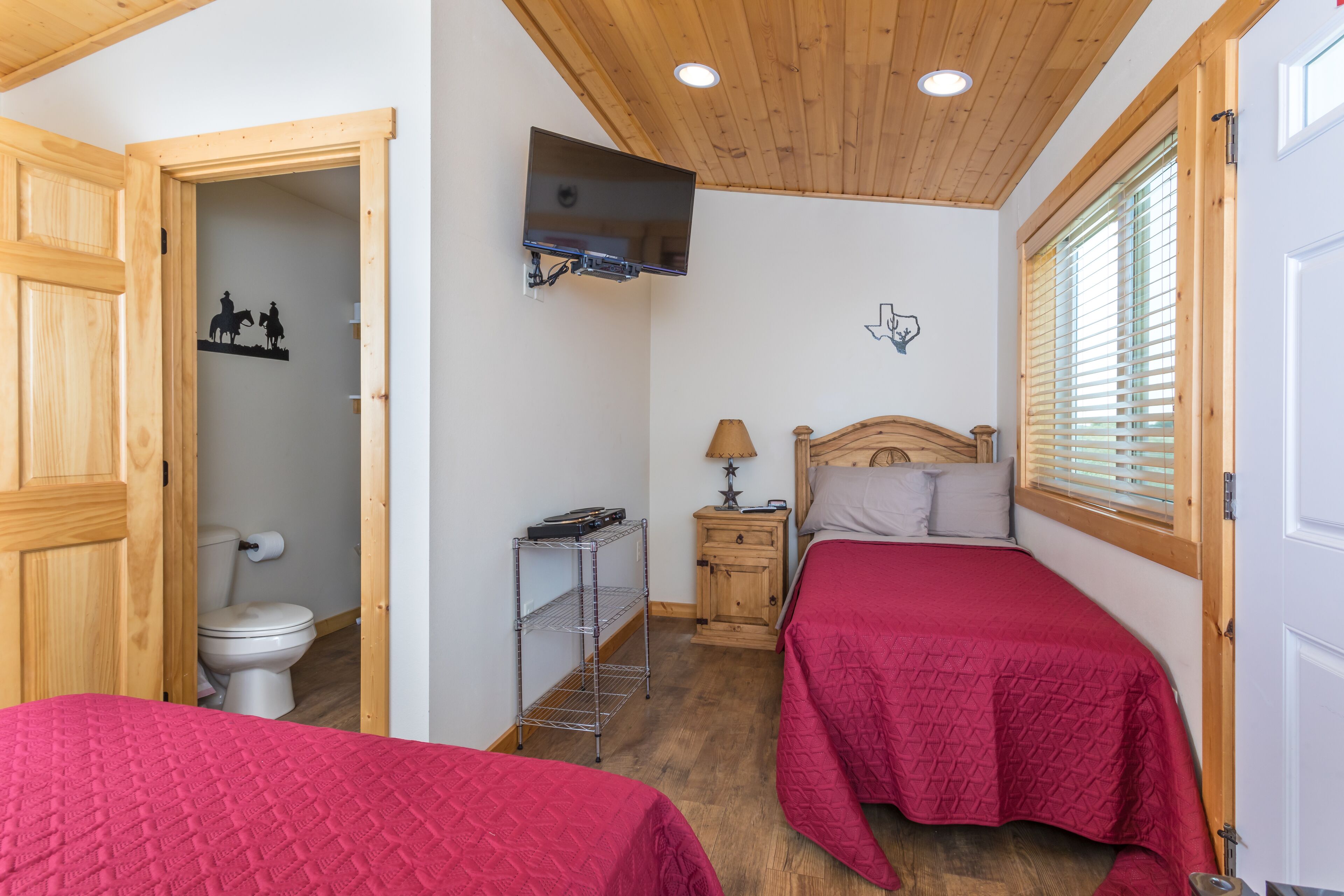 Quarto com duas camas, teto de madeira, televisão fixada na parede, porta de madeira e janela com persianas.
