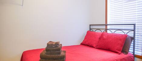 Una cama con colcha y almohadas rojas, una pila de toallas dobladas y una ventana con persianas.