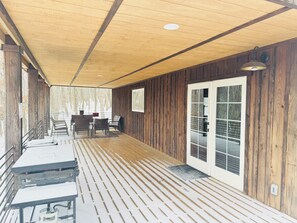 Um espaço coberto ao ar livre com piso de madeira, área de estar com cadeiras e mesas, e uma porta que dá para um espaço externo.
