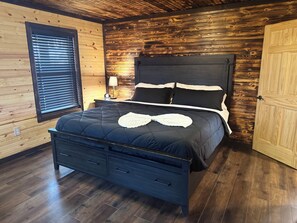 Quarto com painéis de madeira, cama preta com almofadas brancas, mesa de cabeceira com abajur e piso de madeira.