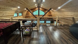A spacious loft-style room with wooden floors and walls, a high ceiling with a fan, and a large window providing a view of greenery outside. - Beautiful Cabin on 20 Acres in Hocking Hills Ohio - Early Bird Discounts! (Rockbridge)
