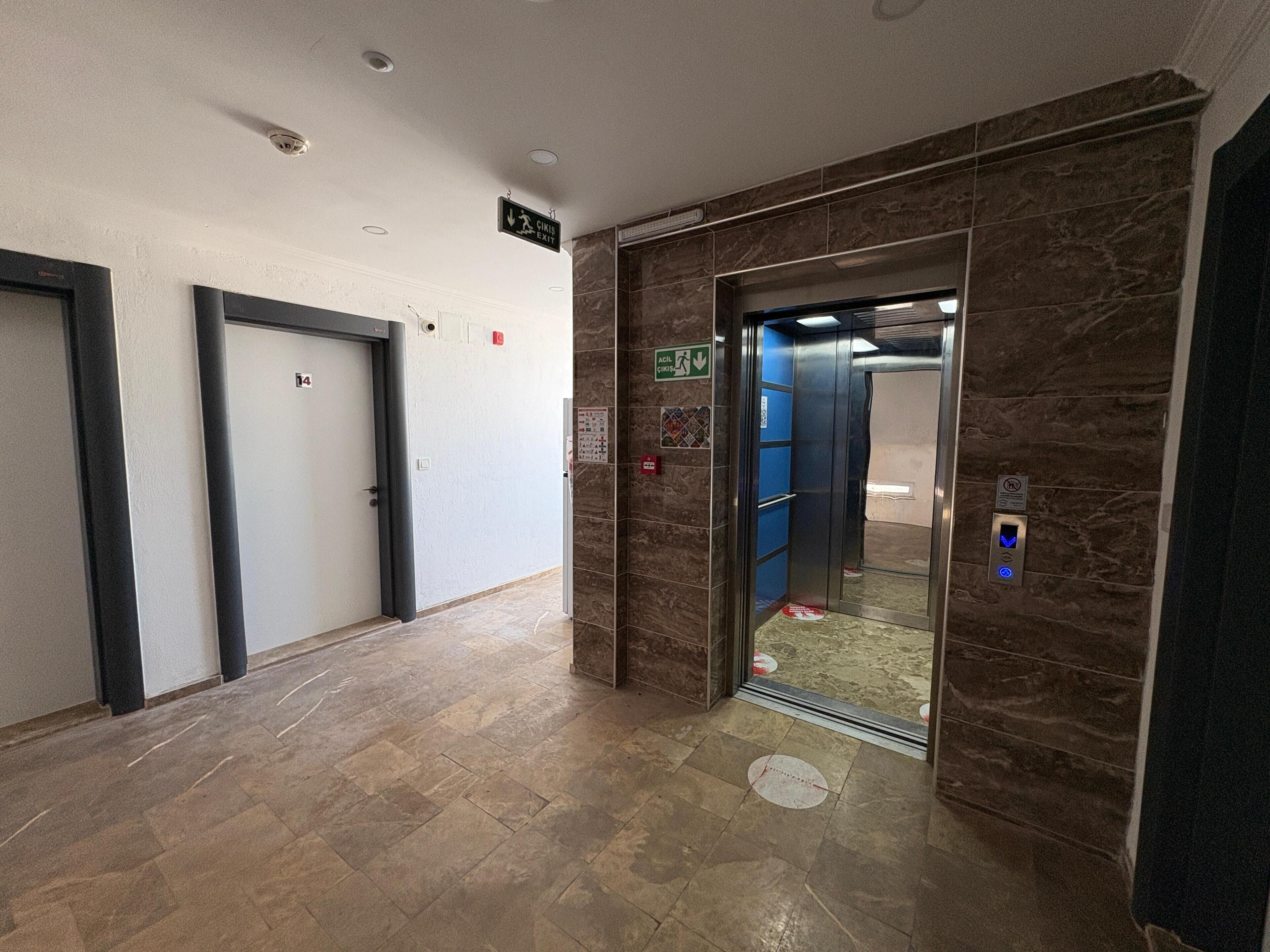 An elevator with a control panel and emergency exit sign, a door with a number sign, and a tiled floor.