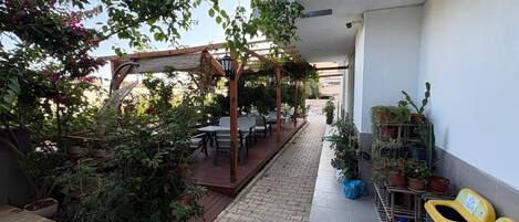 Un patio techado al aire libre con mesas y sillas de madera, rodeado de plantas en macetas y una pérgola con luces colgantes.