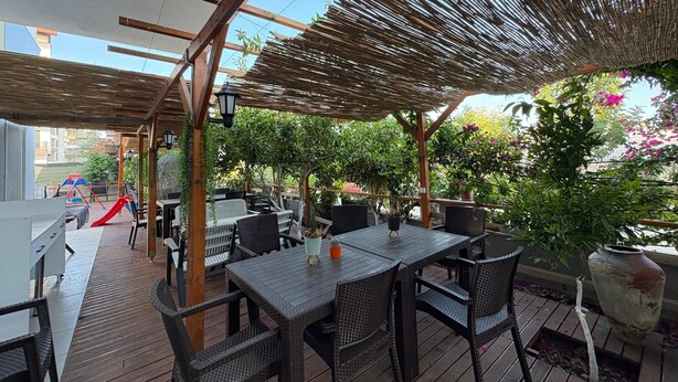 Un patio al aire libre con mesas y sillas de madera, una zona de juegos para niños y plantas en macetas.