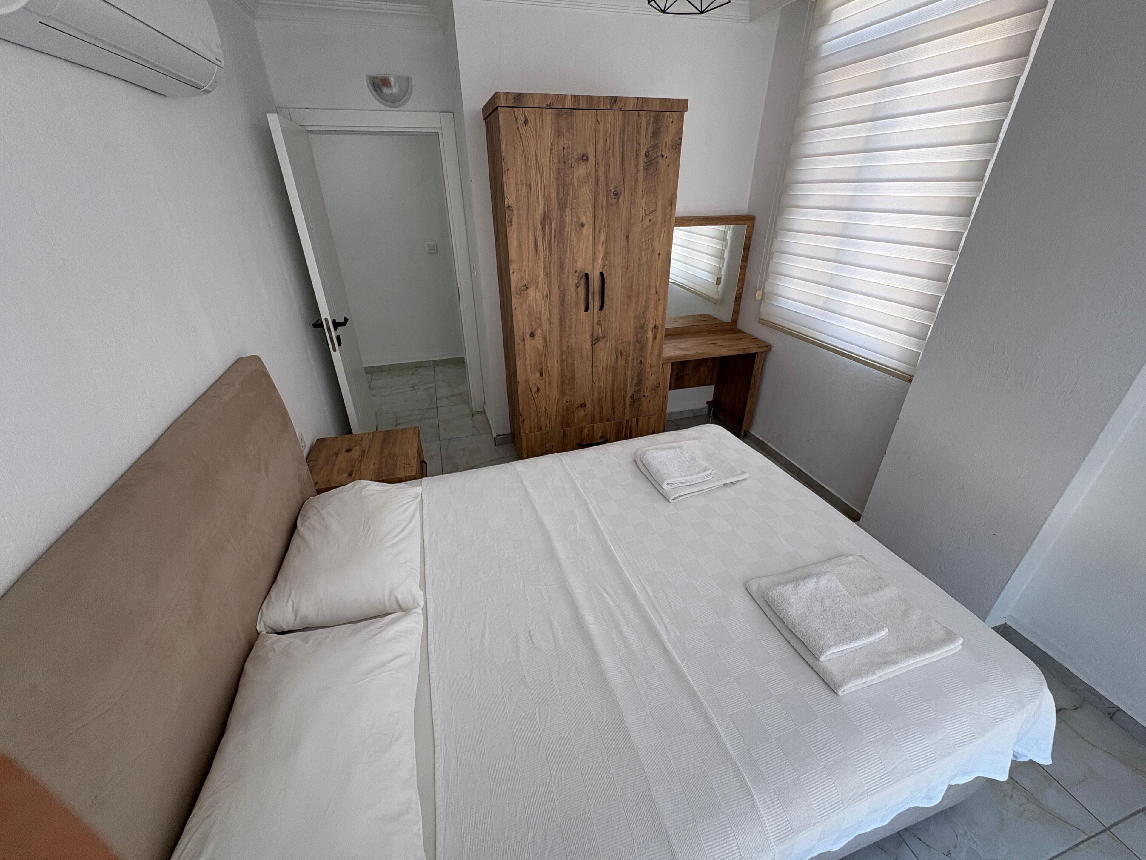 Ein Schlafzimmer mit einem Bett, einem Holzschrank, einem kleinen Holztisch und einem Fenster mit Jalousien.