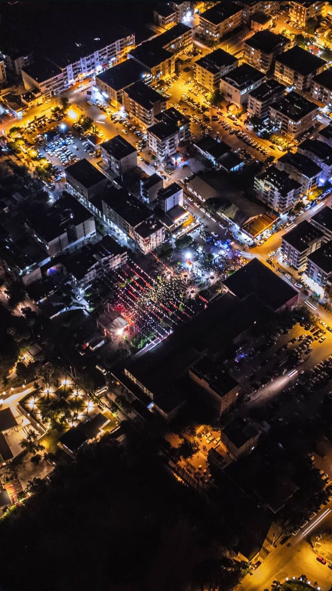 Vista aérea de un paisaje urbano nocturno con edificios iluminados, calles y una gran concentración de gente.