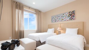 Een hotelkamer met twee bedden, een raam met gordijnen, een speelgoed tractor op een tafel en een kleurrijk kunstwerk aan de muur.