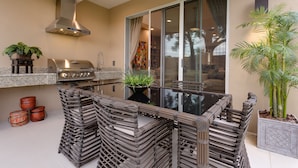 Een buitenkeuken met een barbecue, een spoelbak en een eethoek met een glazen tafel en stoelen.