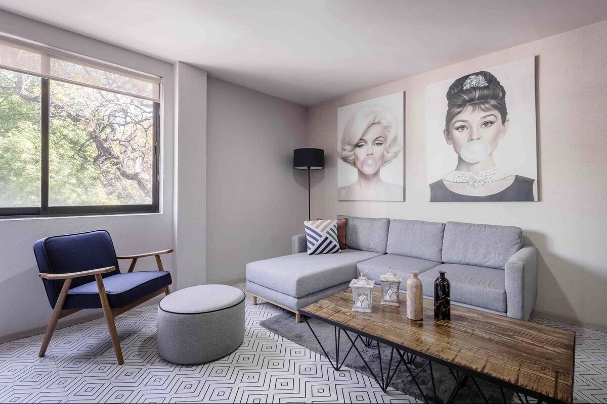 A modern living room with a grey sofa, a blue armchair, a coffee table, and a floor lamp. There are two wall-mounted artworks and a window with a view of trees.