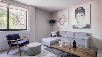Un moderno salón con un sofá gris, un sillón azul, una mesa de centro y una lámpara de pie. Se observan dos obras de arte colgadas en la pared y una ventana con vistas a árboles.