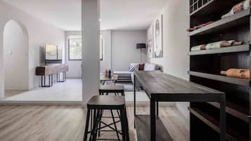 Una sala de estar moderna con una larga mesa de madera, dos taburetes altos, un sofá y un televisor. Se observa una estantería con objetos decorativos y una ventana que proporciona luz natural.