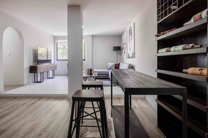 Ein modernes Wohnzimmer mit einem langen Holztisch, zwei Barhocker, einer Couch und einem Fernseher. Ein Bücherregal mit Dekorationselementen und ein Fenster sorgen für natürliches Licht.