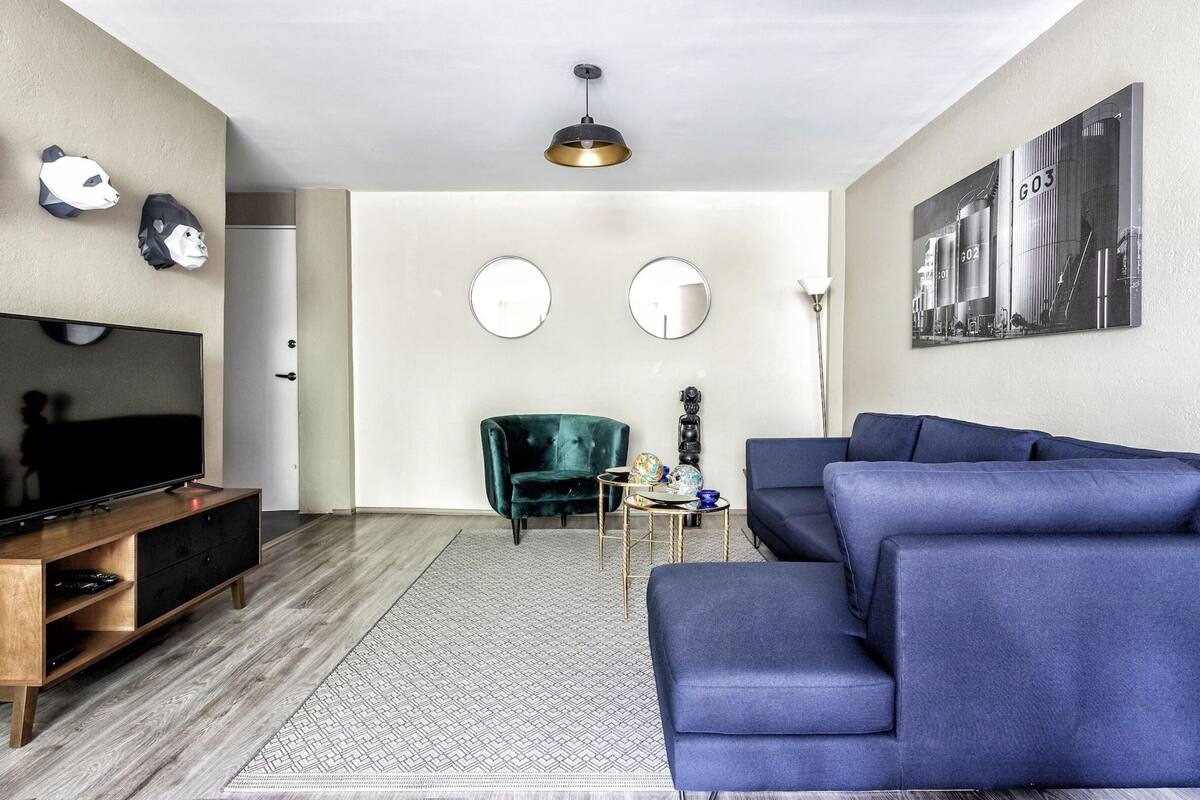 A modern living room with a blue sofa, a wooden entertainment unit, a green armchair, and a large mirror on the wall.