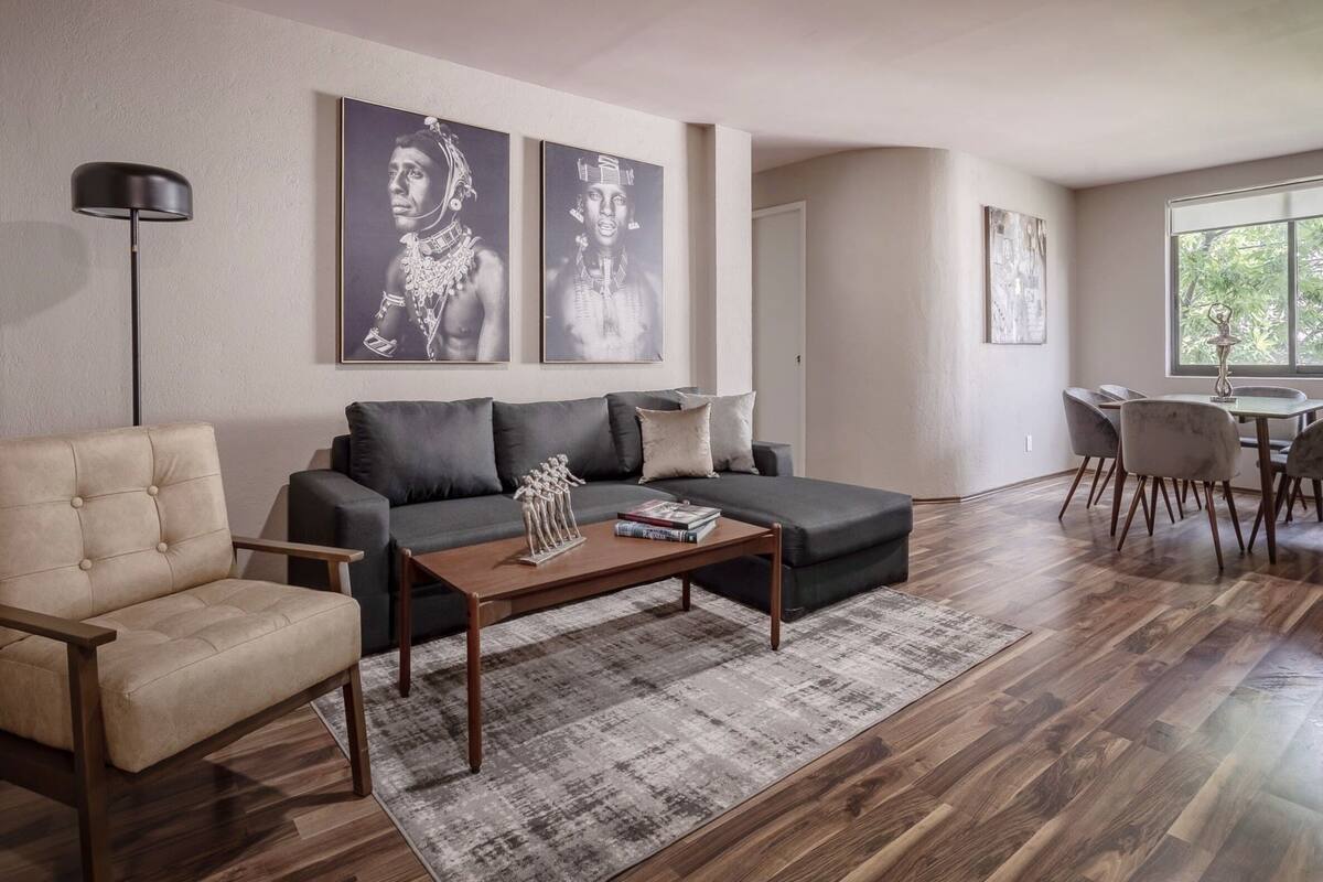 A modern living room with a beige sofa, a dark grey sofa, a wooden coffee table, and a dining area with a table and chairs. There are two framed artworks on the wall.