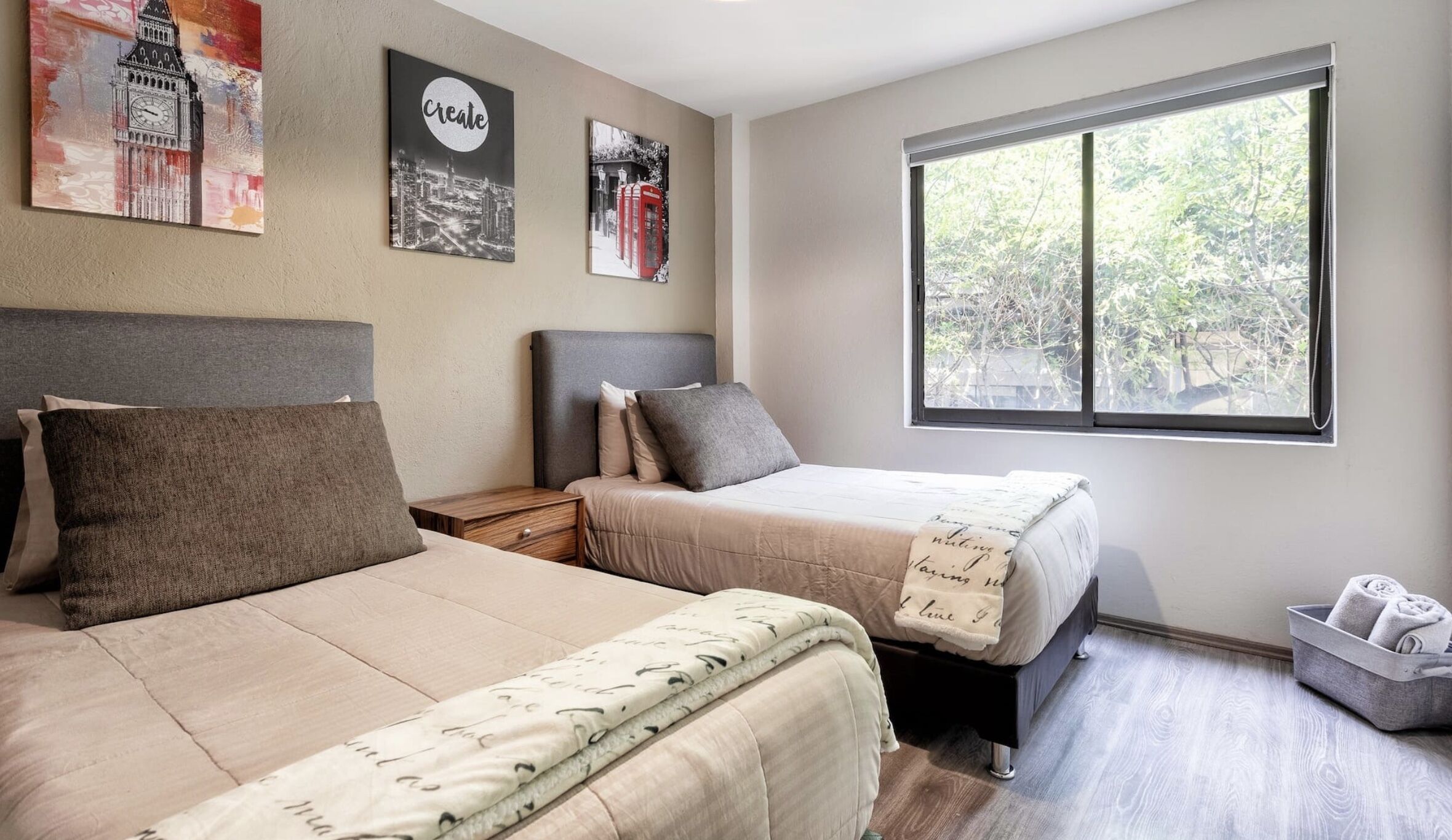 A bedroom with two beds, a nightstand, a window with a view of trees, and wall art including a clock and a 'create' sign.