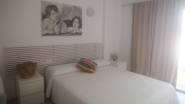 Habitación de hotel con cama, cabecero, mesita de noche, cesta, almohada colorida, un cuadro en la pared y una ventana con cortinas.