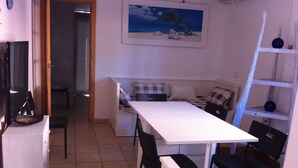Una habitación pequeña con una mesa blanca, dos sillas negras, una cama con ropa de cama a cuadros, un cuadro en la pared y una escalera apoyada en la pared.