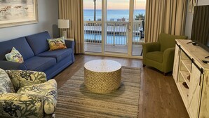 Een woonkamer met een blauwe bank, een groene fauteuil, een ronde salontafel en een tv-meubel. Een groot raam biedt uitzicht op de oceaan en een palmboom.