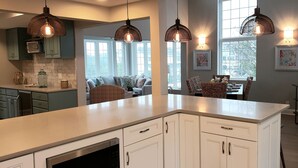 Une cuisine moderne avec des meubles blancs, un grand îlot central, des luminaires suspendus et un coin repas avec des meubles en osier.