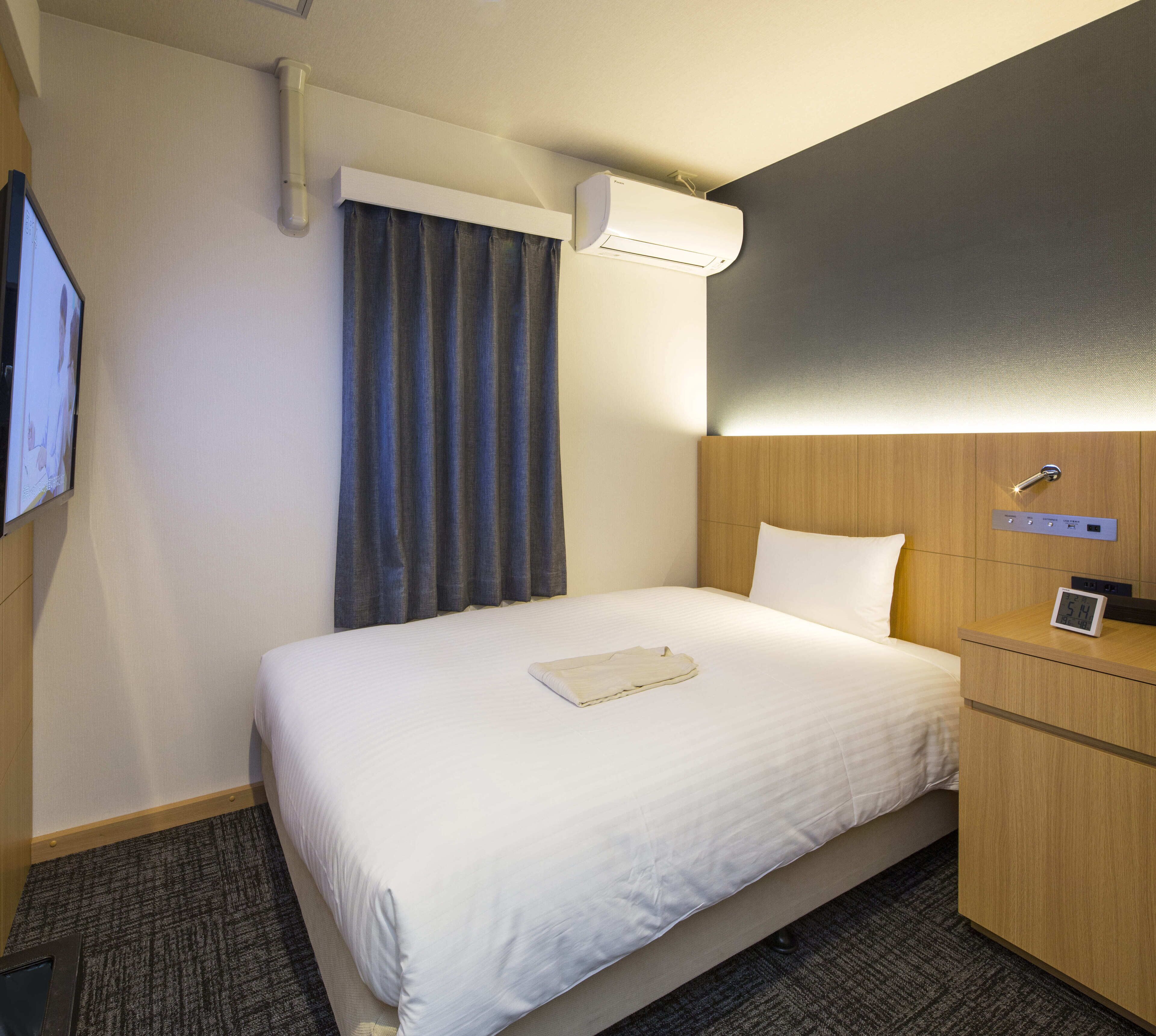 Une chambre d’hôtel avec un lit, une table de chevet avec une horloge, un climatiseur fixé au mur et un téléviseur mural.
