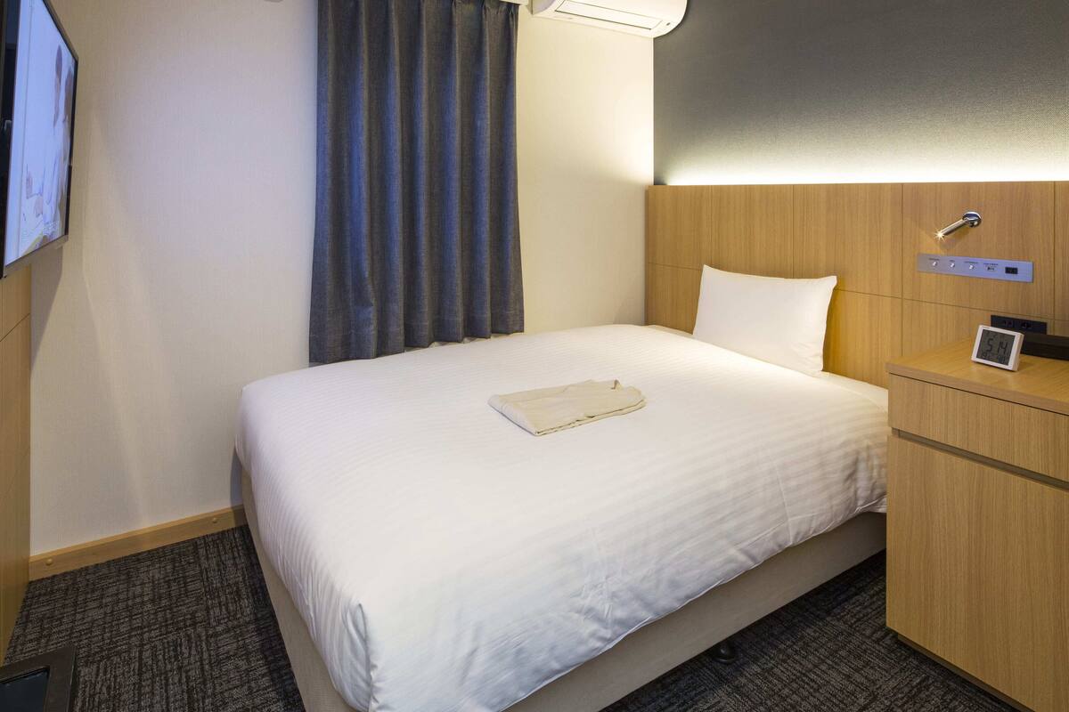 Une chambre d’hôtel avec un lit, une table de chevet avec une horloge, un climatiseur fixé au mur et un téléviseur mural.