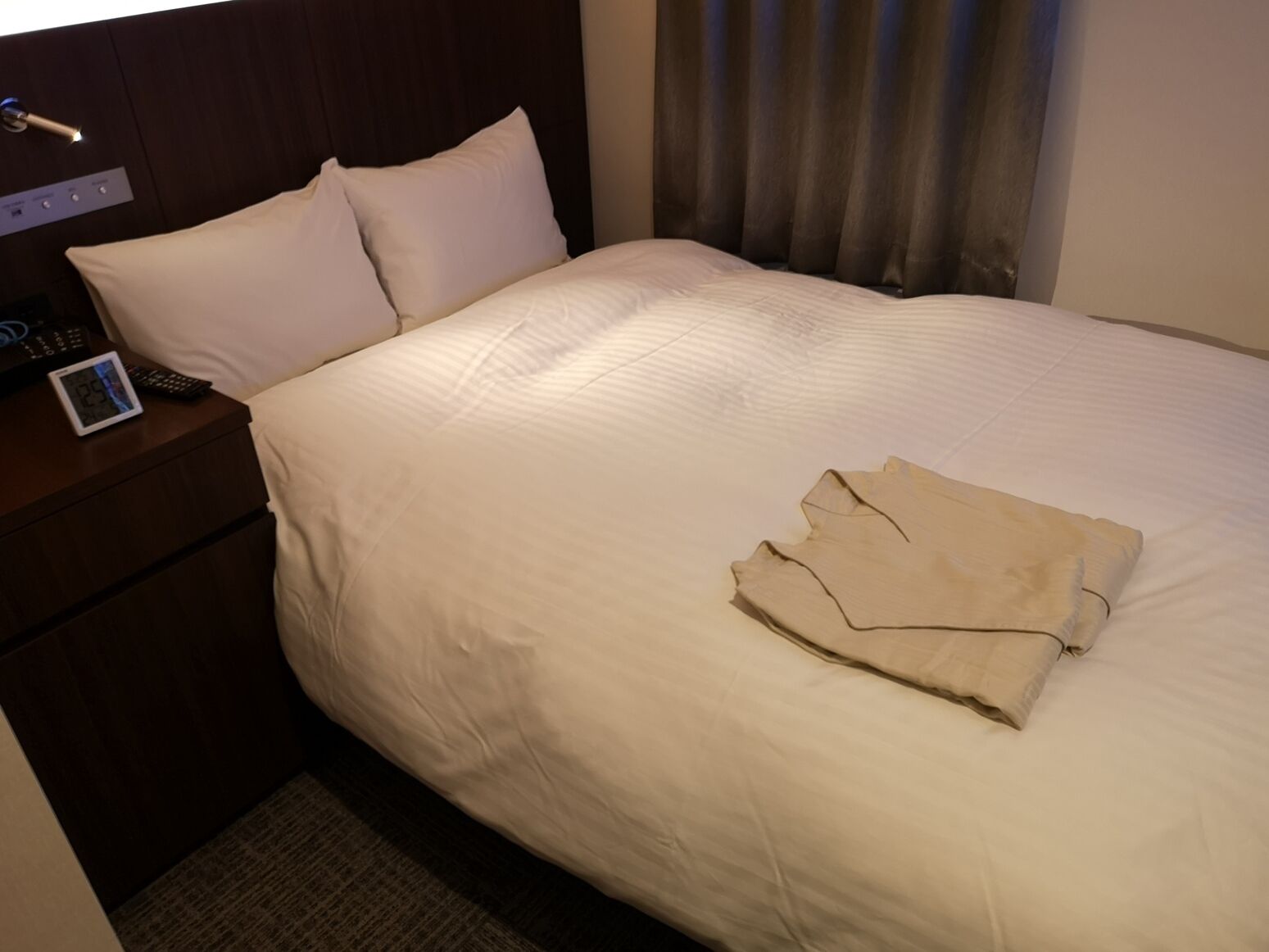Ein Hotelzimmer mit einem ordentlich bezogenen Bett, einem Nachttisch mit Lampe und einer digitalen Uhr.