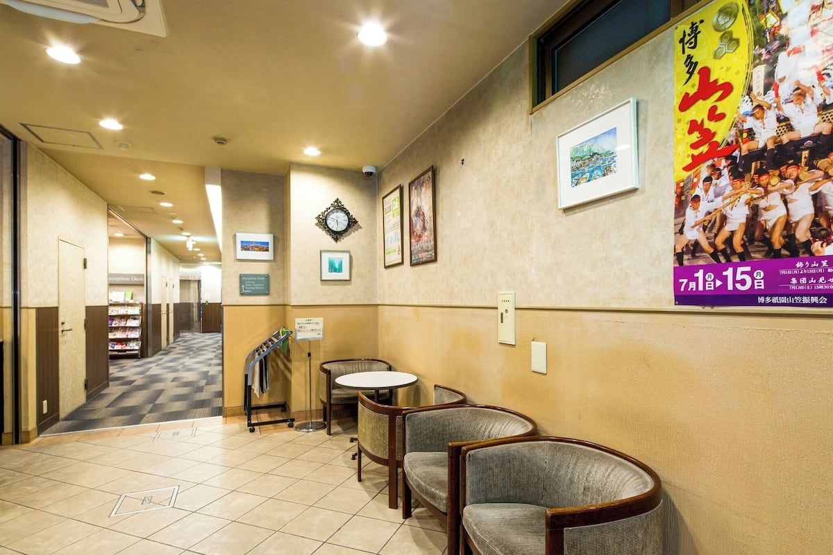 Un hall d’hôtel avec un comptoir d’accueil, des chaises, une horloge, des tableaux encadrés et une affiche accrochée au mur.