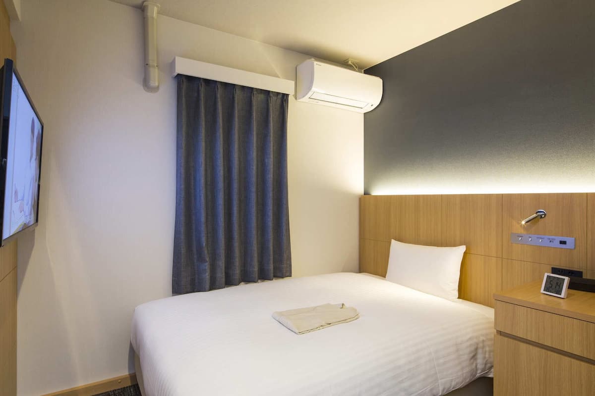 Une chambre d’hôtel comprenant un lit, une table de chevet avec une horloge, un appareil de climatisation, une télévision et une fenêtre avec des rideaux.