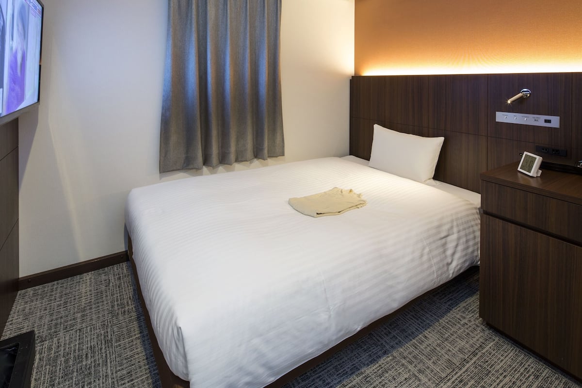 Une chambre d’hôtel équipée d’un lit, d’une télévision, d’un climatiseur, d’une table de chevet avec une lampe et d’un panneau de commande fixé au mur.