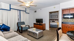 A living room with a sofa, armchair, ottoman, TV, ceiling fan, and kitchen area with cabinets and microwave.