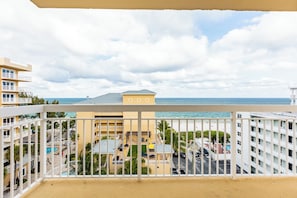A balcony with a view of a beach, a pool, and a building with multiple floors.