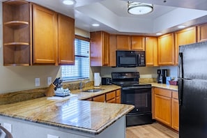 A kitchen with wooden cabinets, a granite countertop, a black microwave, a stove, and a refrigerator. - Pompano Beach | Couples Ocean Coast Retreat | Pool + Beach (Pompano Beach)