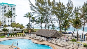 Área de piscina en un resort, con sillas de descanso, una cabaña con techo de paja y vista al océano.