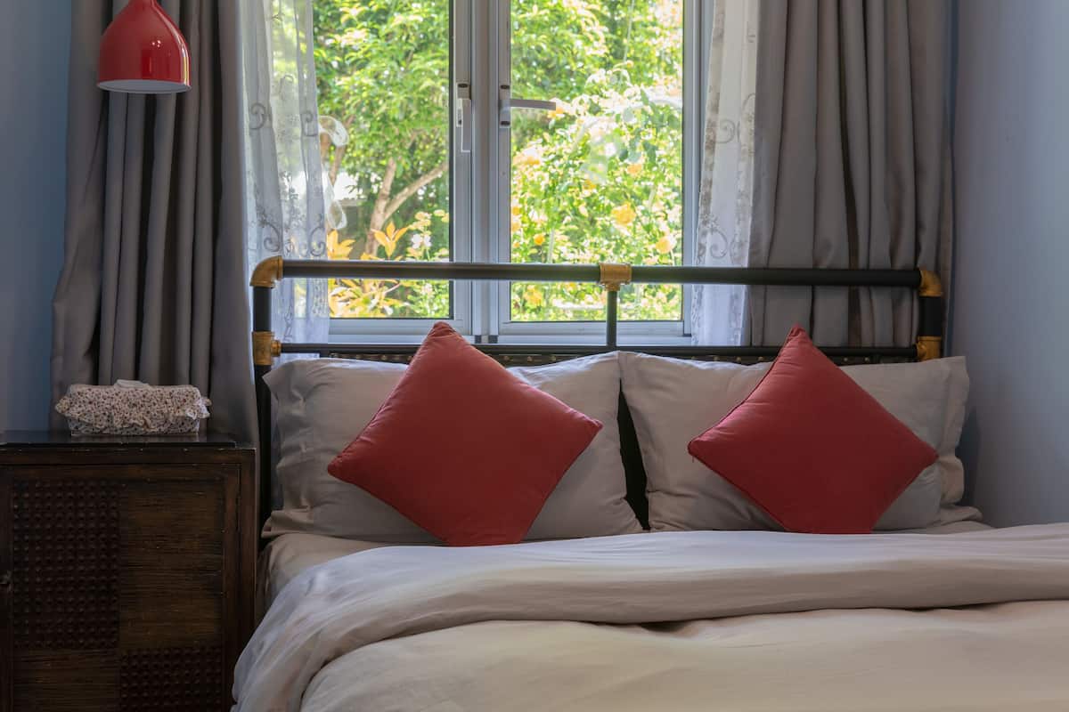 A bed with white and red pillows, a wooden nightstand, a red lamp, and a window with curtains.