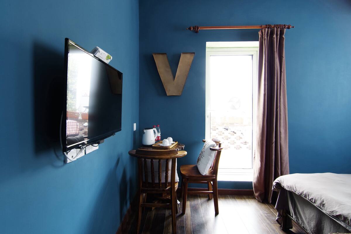 A room with a TV mounted on the wall, a large V-shaped decoration, a window with curtains, a bed, and a small table with a kettle and cups.