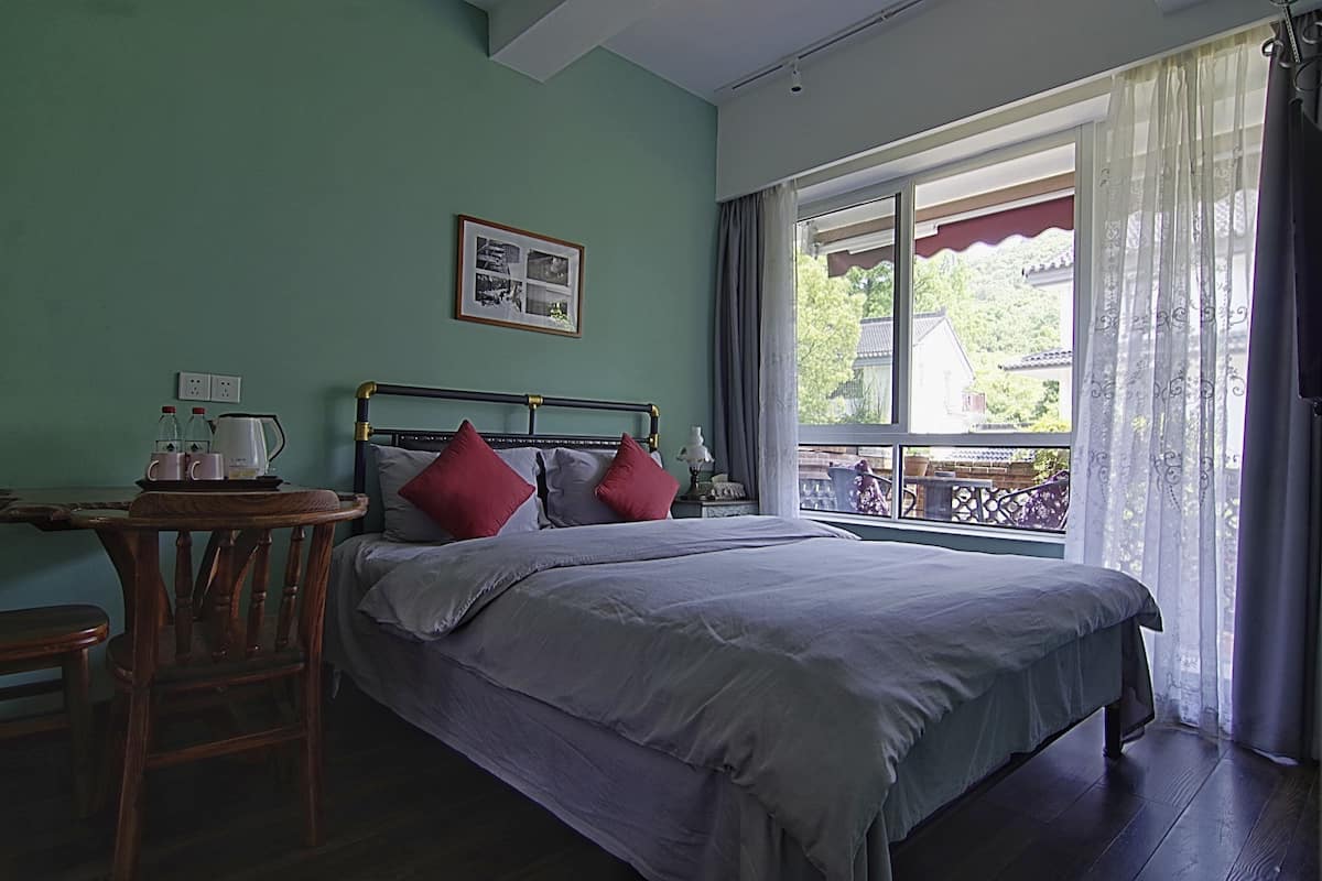 A bedroom with a bed, a wooden chair, a table with a kettle, and a window with curtains.