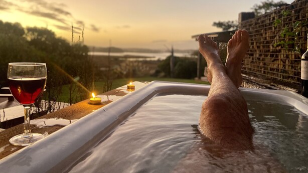 A person relaxing in a hot tub with a glass of red wine and candles on a table, overlooking a scenic view.