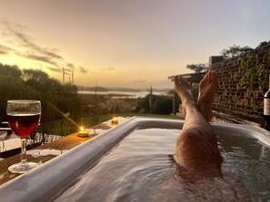 Une personne se détend dans un bain à remous, un verre de vin rouge à la main, avec des bougies sur une table, et profite d’une vue panoramique.