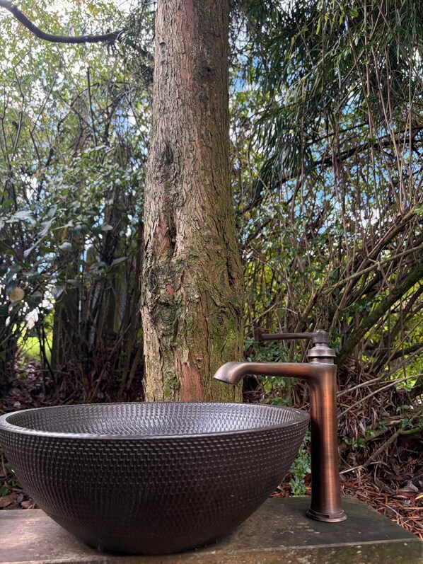 A large, textured bowl sink with a single faucet, placed outdoors with a tree and foliage in the background.