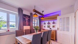 Een eethoek met een houten tafel, stoelen en een bloemenvaas. Er is een raam met gordijnen, een radiator en een deur met ruit.