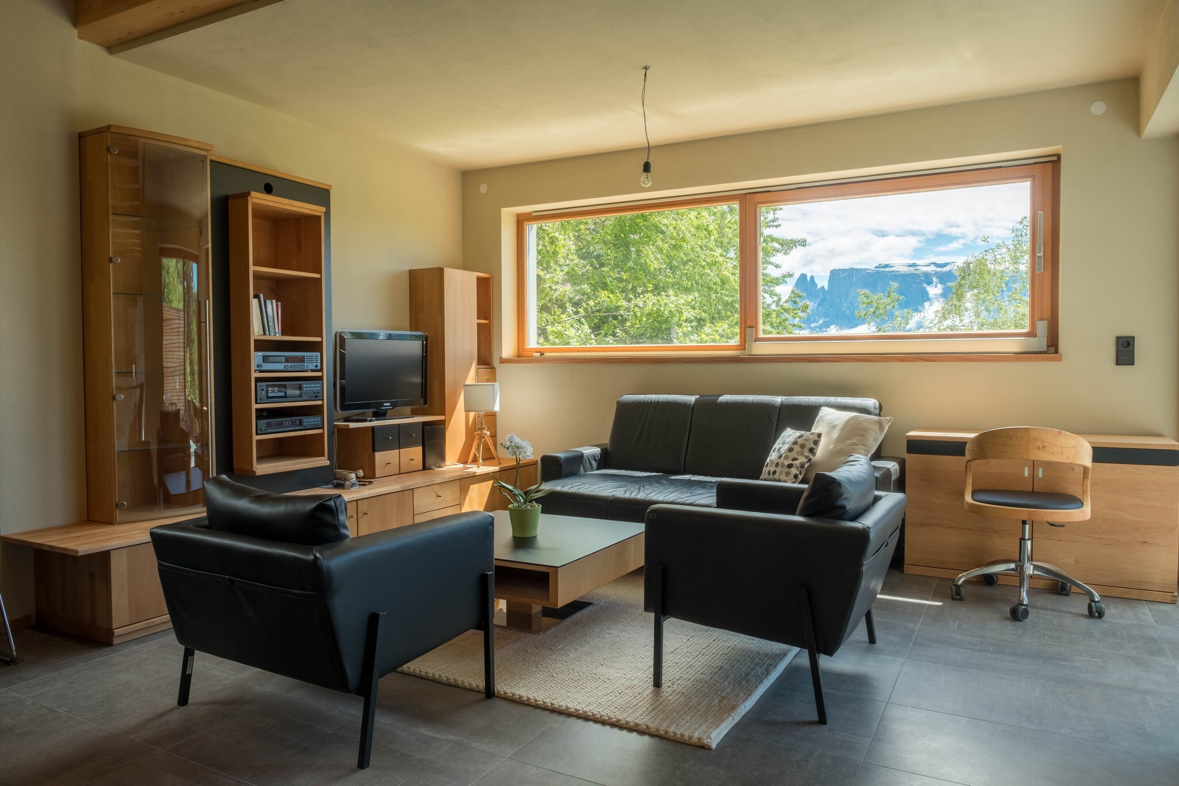 Een moderne woonkamer met een bank, stoelen, een televisie en een groot raam met uitzicht op bomen en bergen.