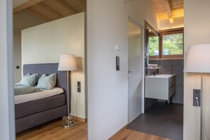 A modern bedroom with a bed, bedside lamp, and a view of a bathroom with a sink and window.