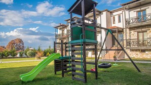 A playground with a green slide and climbing structure, set against a backdrop of a residential area with stone buildings and a clear sky. - Paroraia boutique rooms (Sintiki)